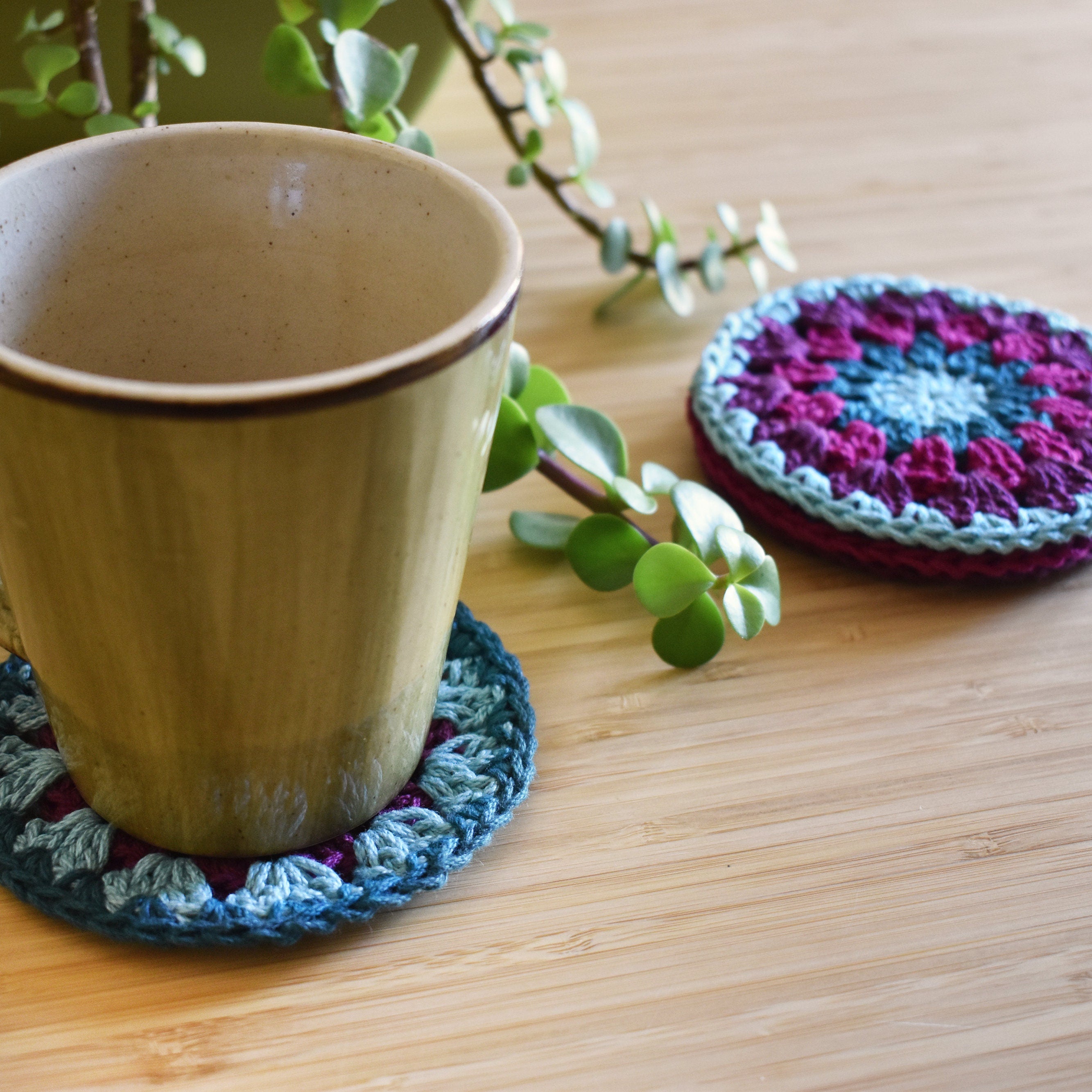 Ceramic mug with a crocheted coaster on a wooden surface, next to another coaster and a plant.