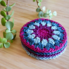 Stack of colorful crocheted coasters on a wooden surface with green plants.