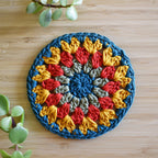 Colorful crocheted round granny sunburst coaster on a wooden surface with green plants.