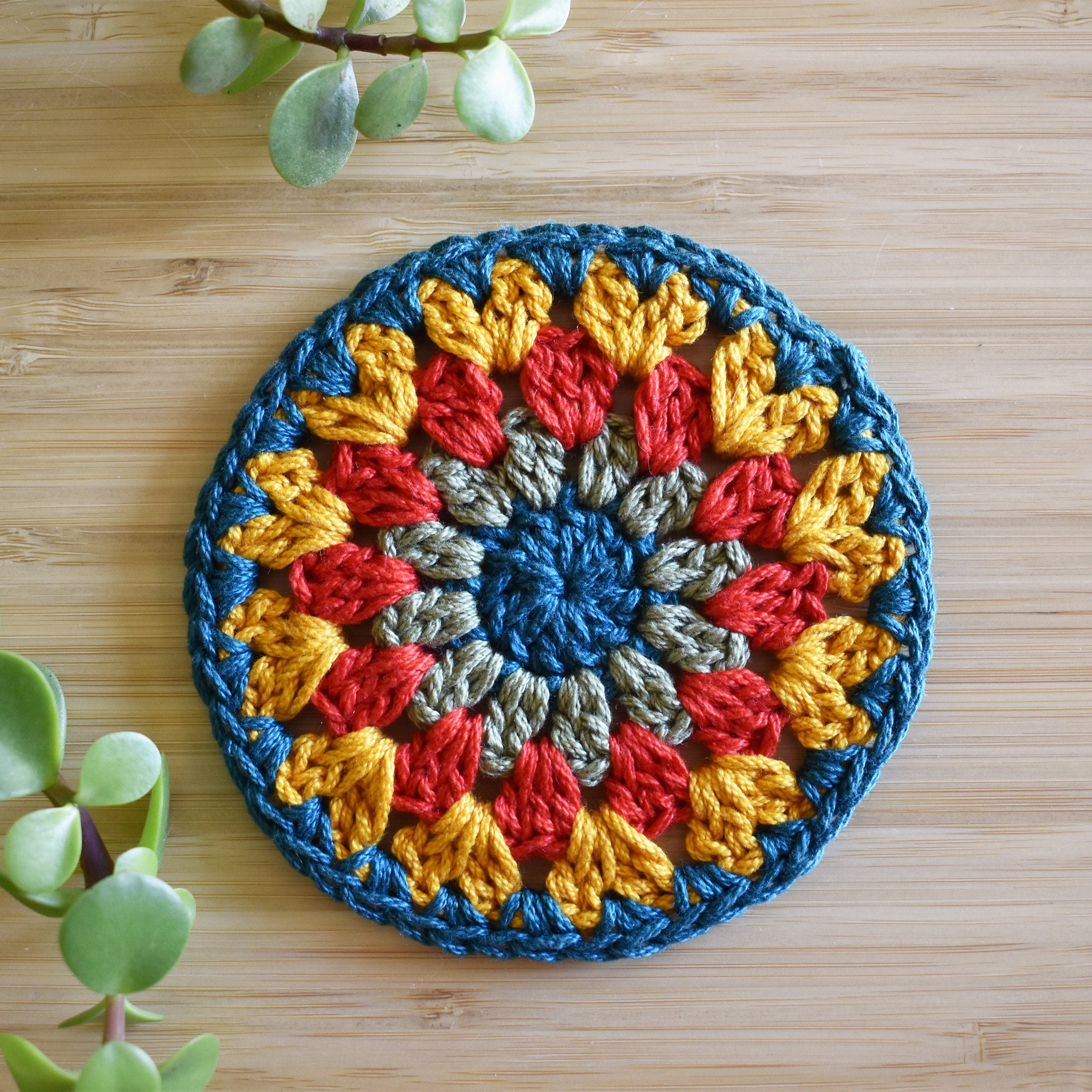 Colorful crocheted round granny sunburst coaster on a wooden surface with green plants.