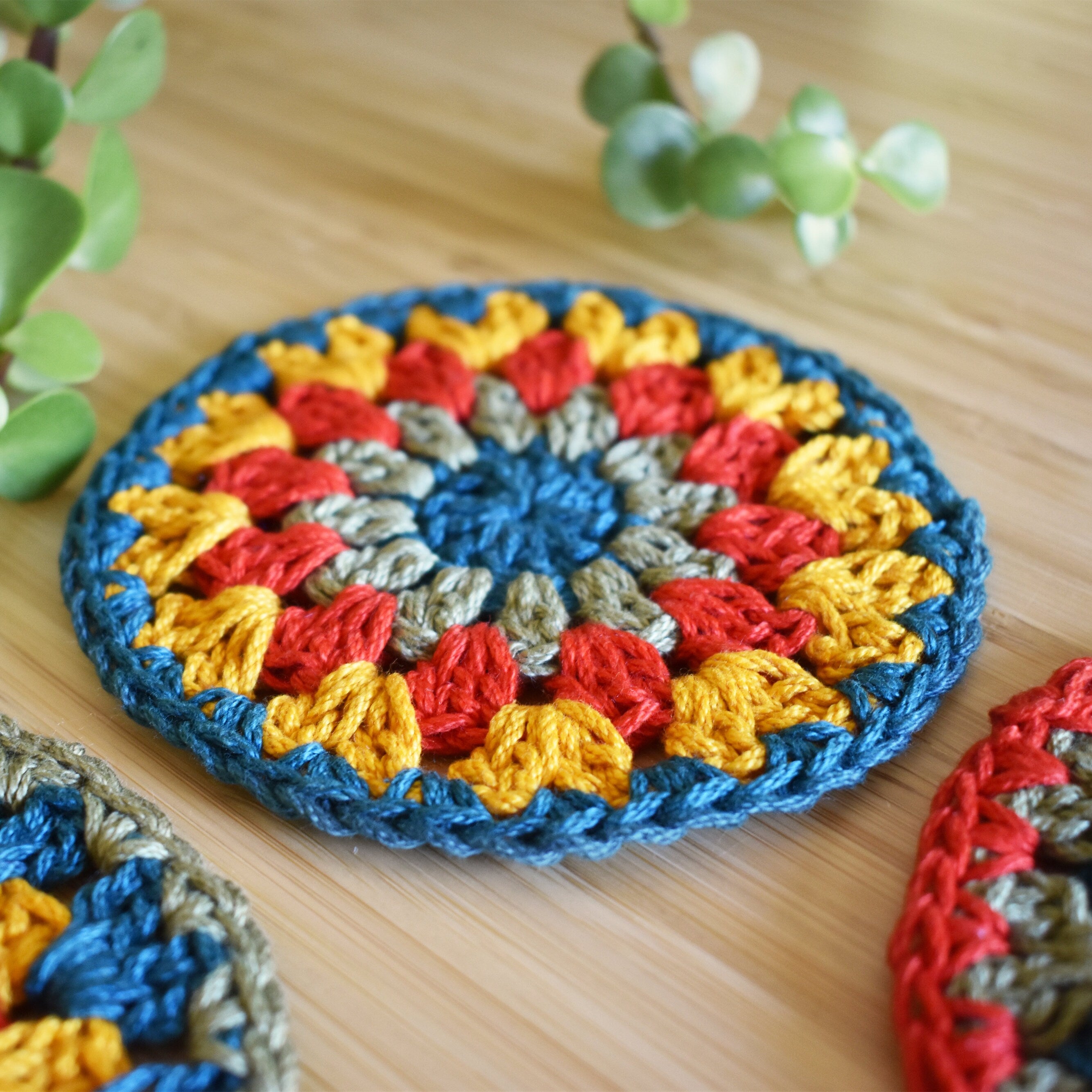 Close-up view of a colorful crocheted coaster on a wooden surface with plants in the background.