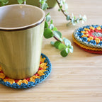 Mug with a colorful crocheted coaster on a wooden surface with a stack of coasters and a plant in the background.