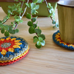 Ceramic mug on a colorful crocheted coaster next to a stack of coasters on a wooden surface with a plant in the background.