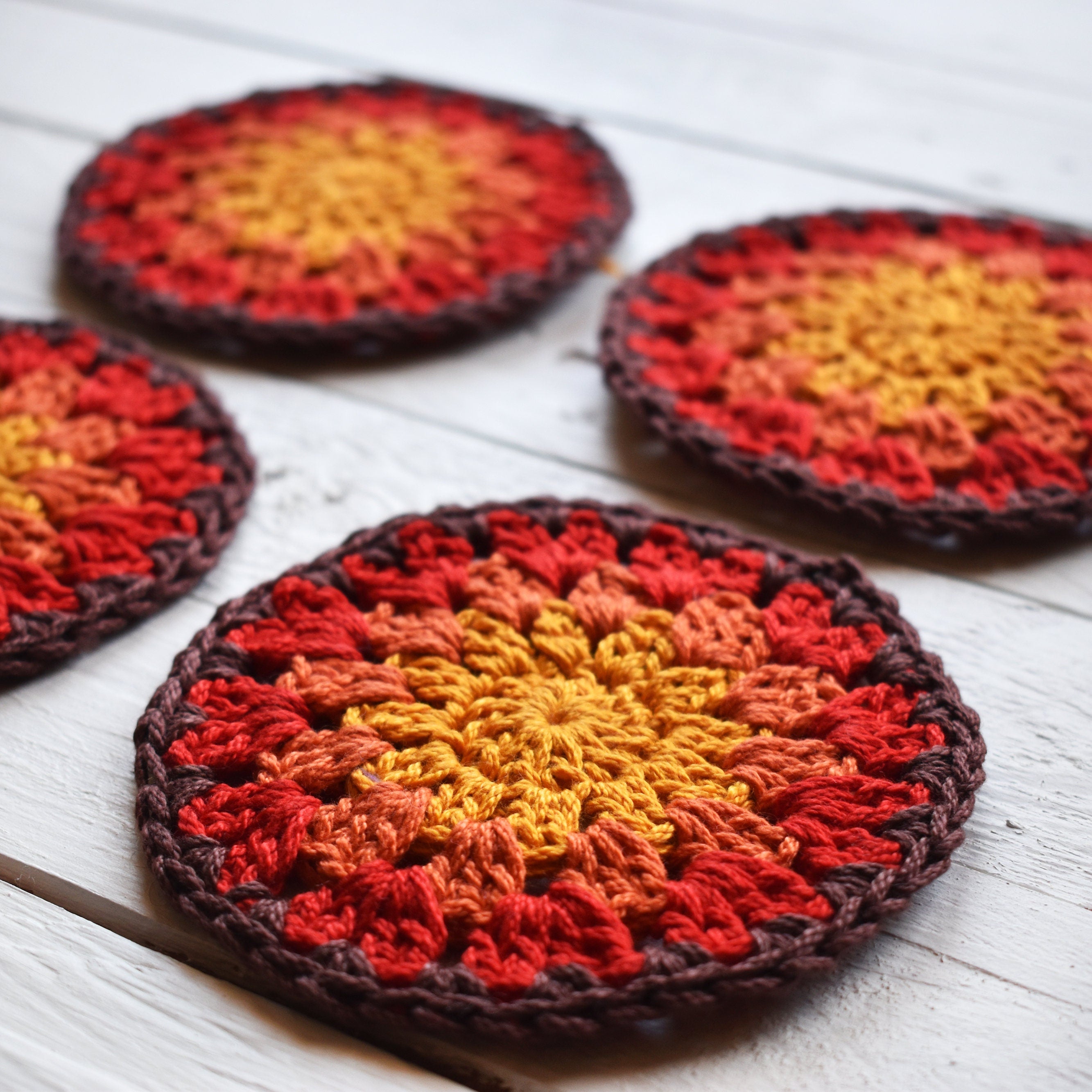 Four crocheted coasters with red, orange, and yellow colors on a white wooden surface.