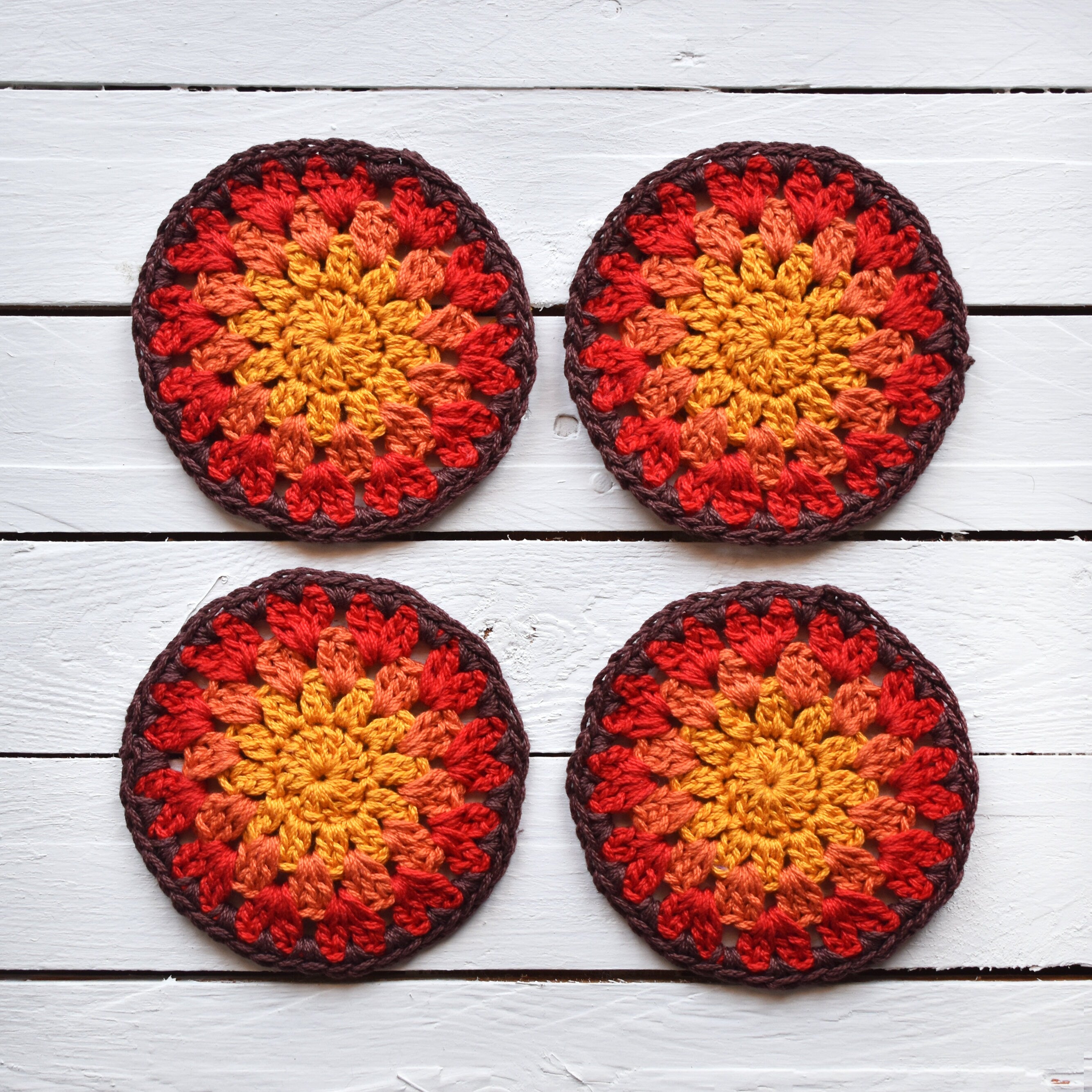 Four crocheted coasters with a granny sunburst design of red, orange, and yellow colors on a white wooden surface.