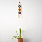 Decorative wall hanging with colorful patterns next to a potted plant on a white background.