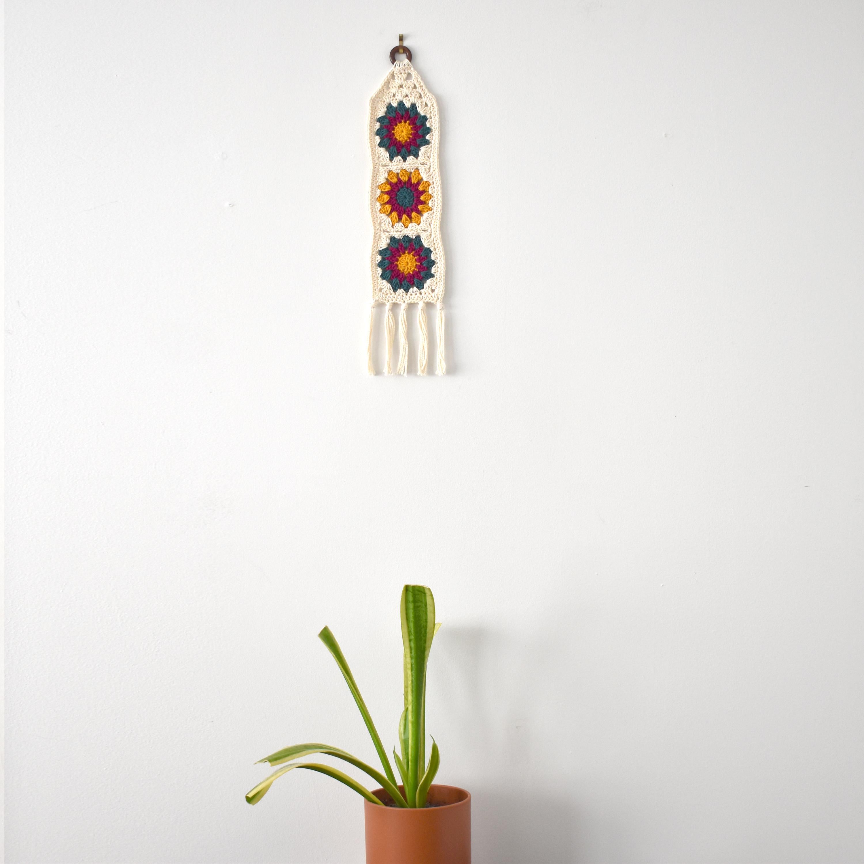 Decorative wall hanging with colorful patterns next to a potted plant on a white background.