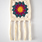 Crocheted wall hanging with colorful flower design and tassels on a white background.