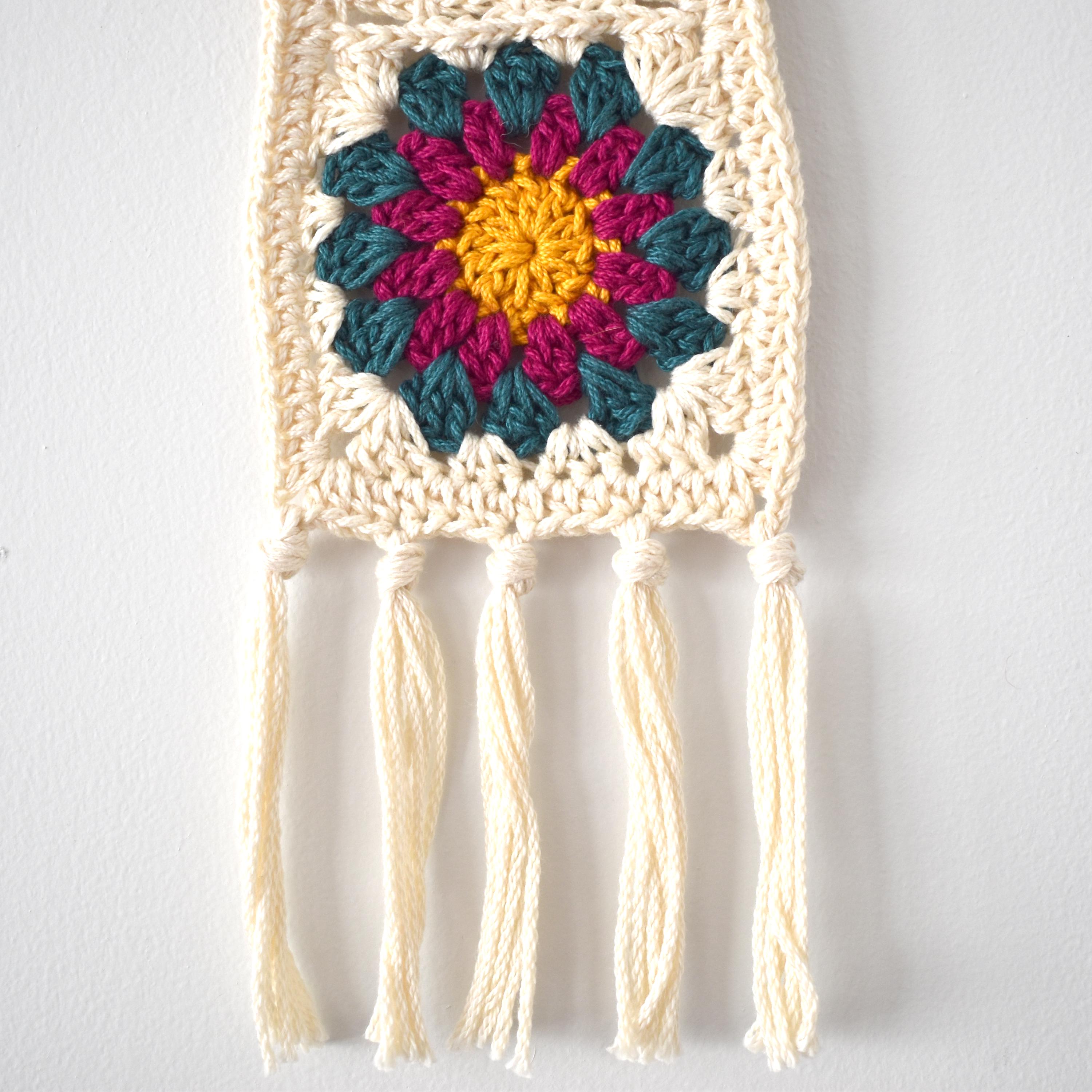 Crocheted wall hanging with colorful flower design and tassels on a white background.
