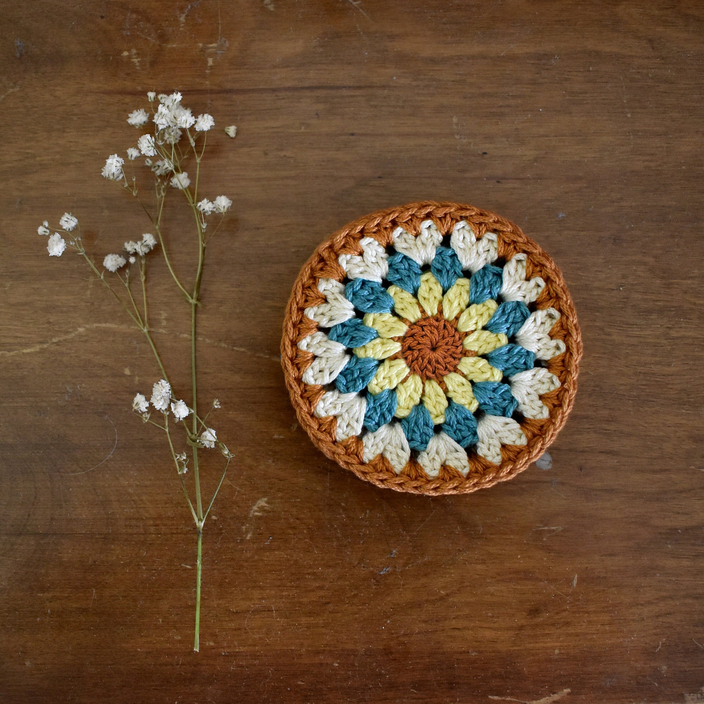 Crocheted coaster with a floral pattern on a wooden surface next to a branch of small white flowers.