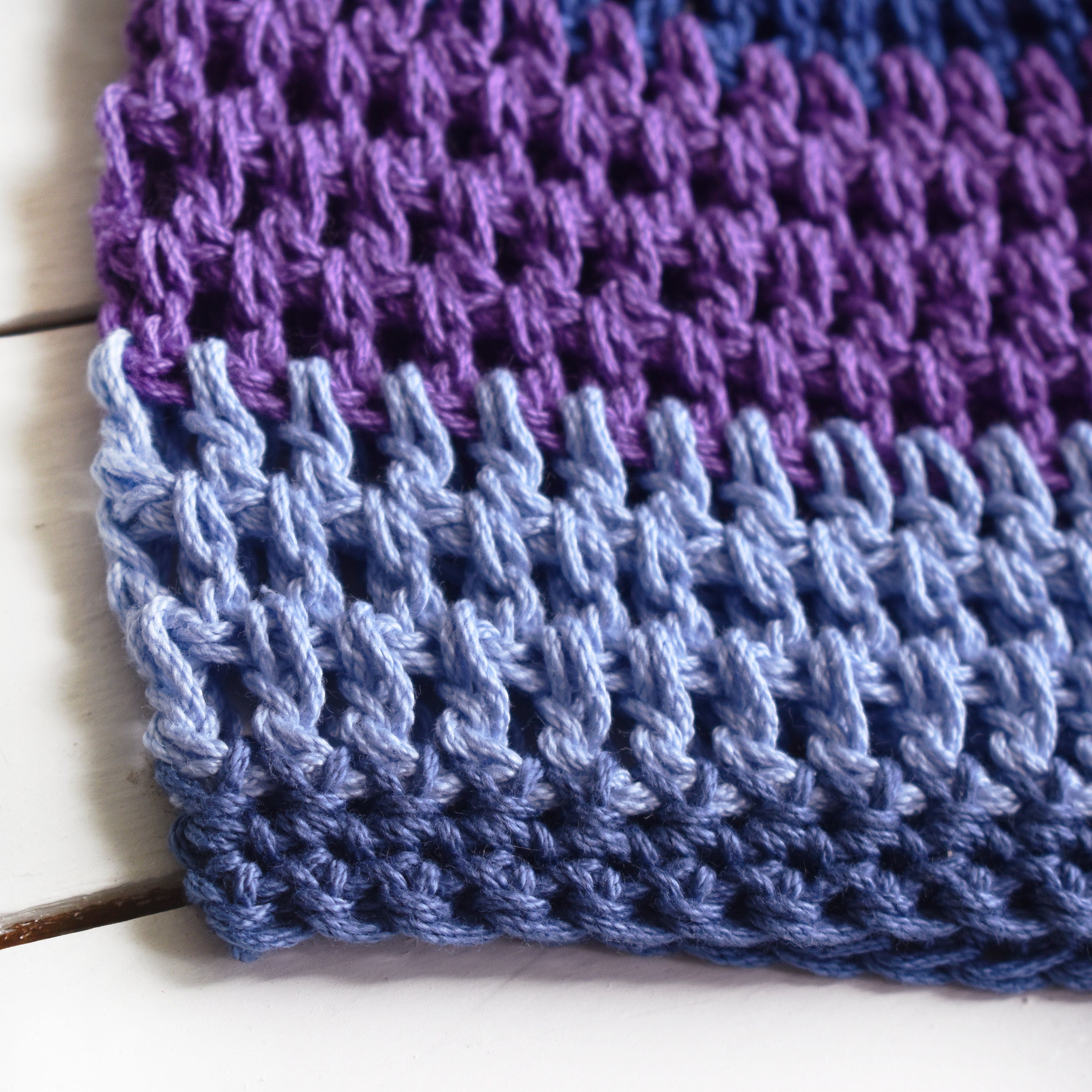 Close-up of a crocheted hat with a purple and blue striped pattern on a white wooden surface.