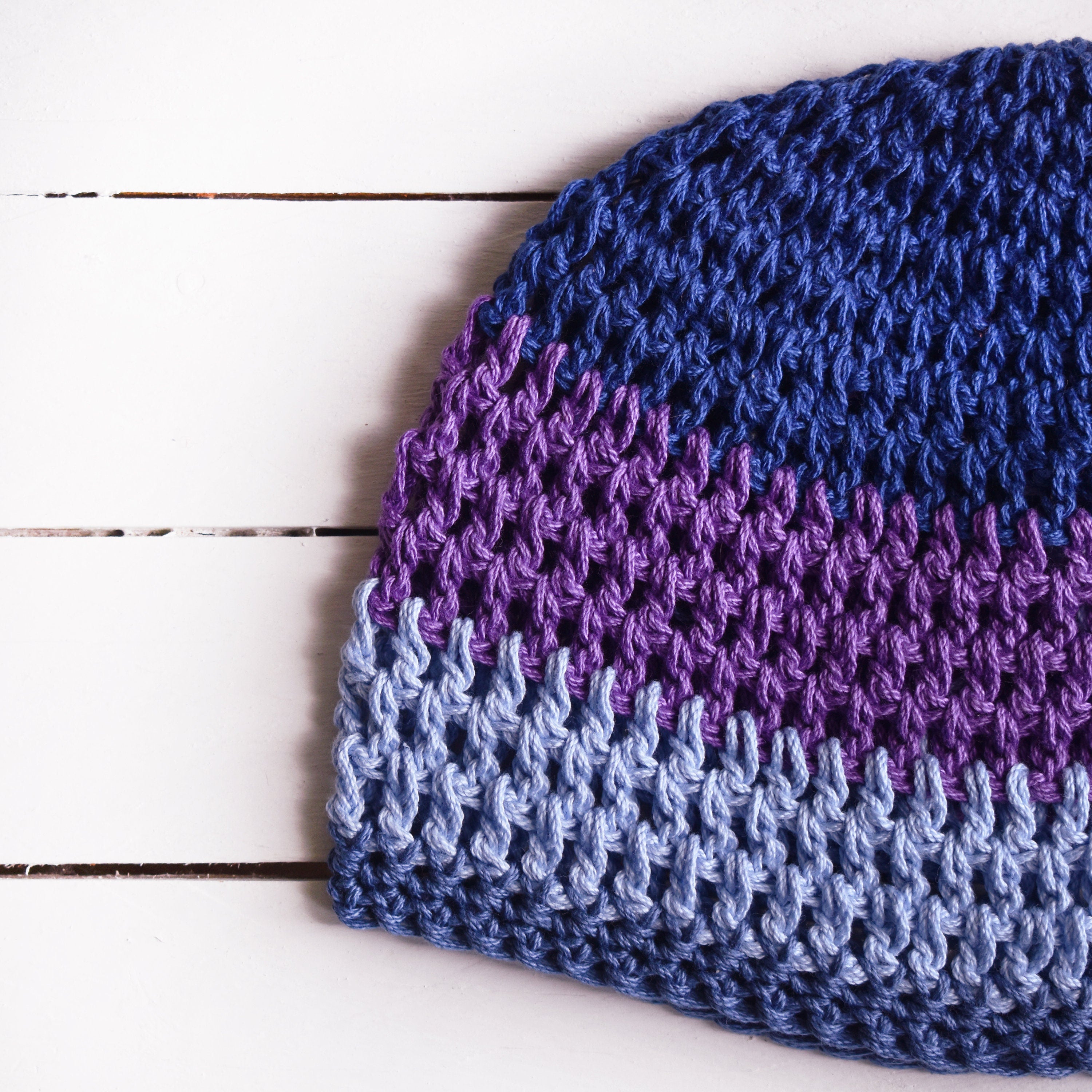 Close-up view of a crocheted beanie with a blue and purple gradient on a white wooden surface.