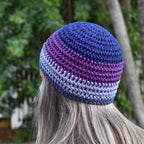 Person wearing a blue-and-purple-striped handmade crochet hat against a blurred natural background.