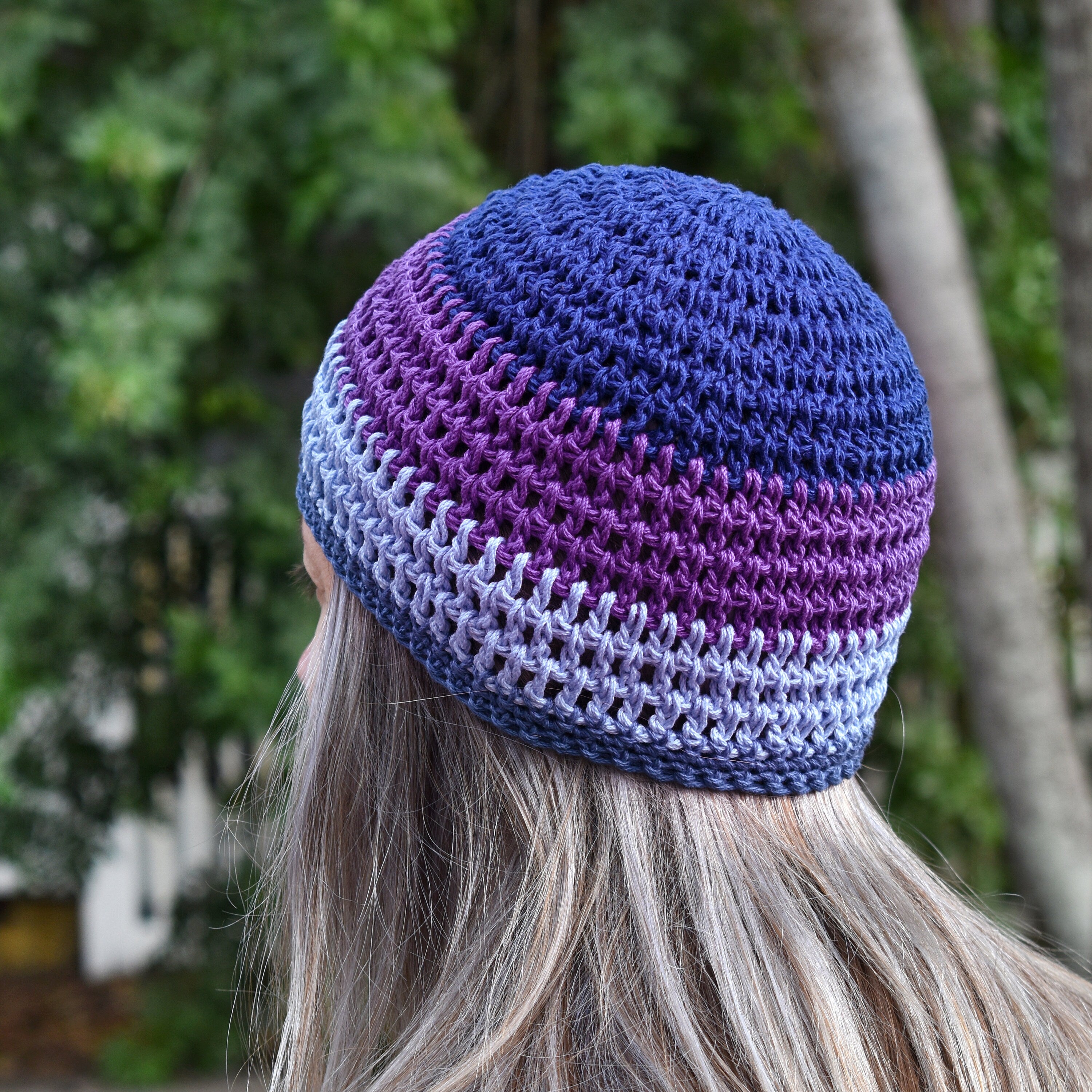 Person wearing a blue-and-purple-striped handmade crochet hat against a blurred natural background.
