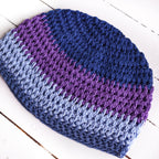 Handmade crochet hat with purple and blue stripes on a white wooden surface.
