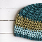 Crocheted beanie with blue and green stripes on a white wooden surface.