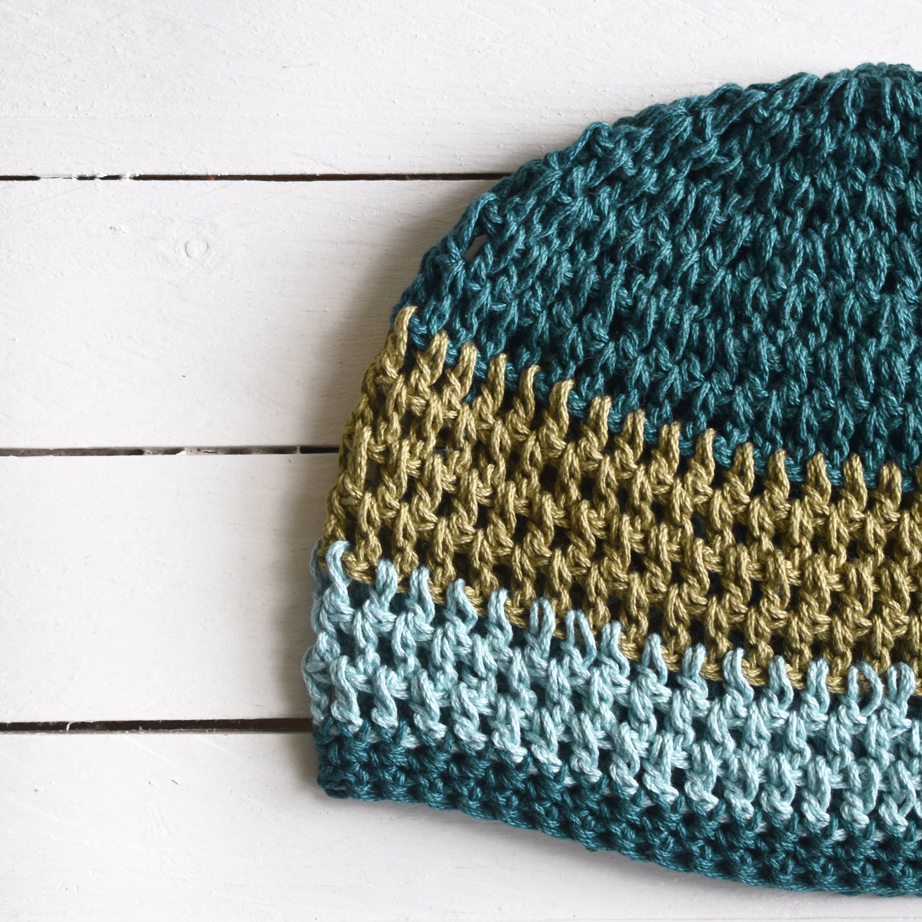 Crocheted beanie with blue and green stripes on a white wooden surface.