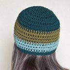 Woman with long hair wearing a crocheted beanie with a teal and green striped pattern against a neutral background.