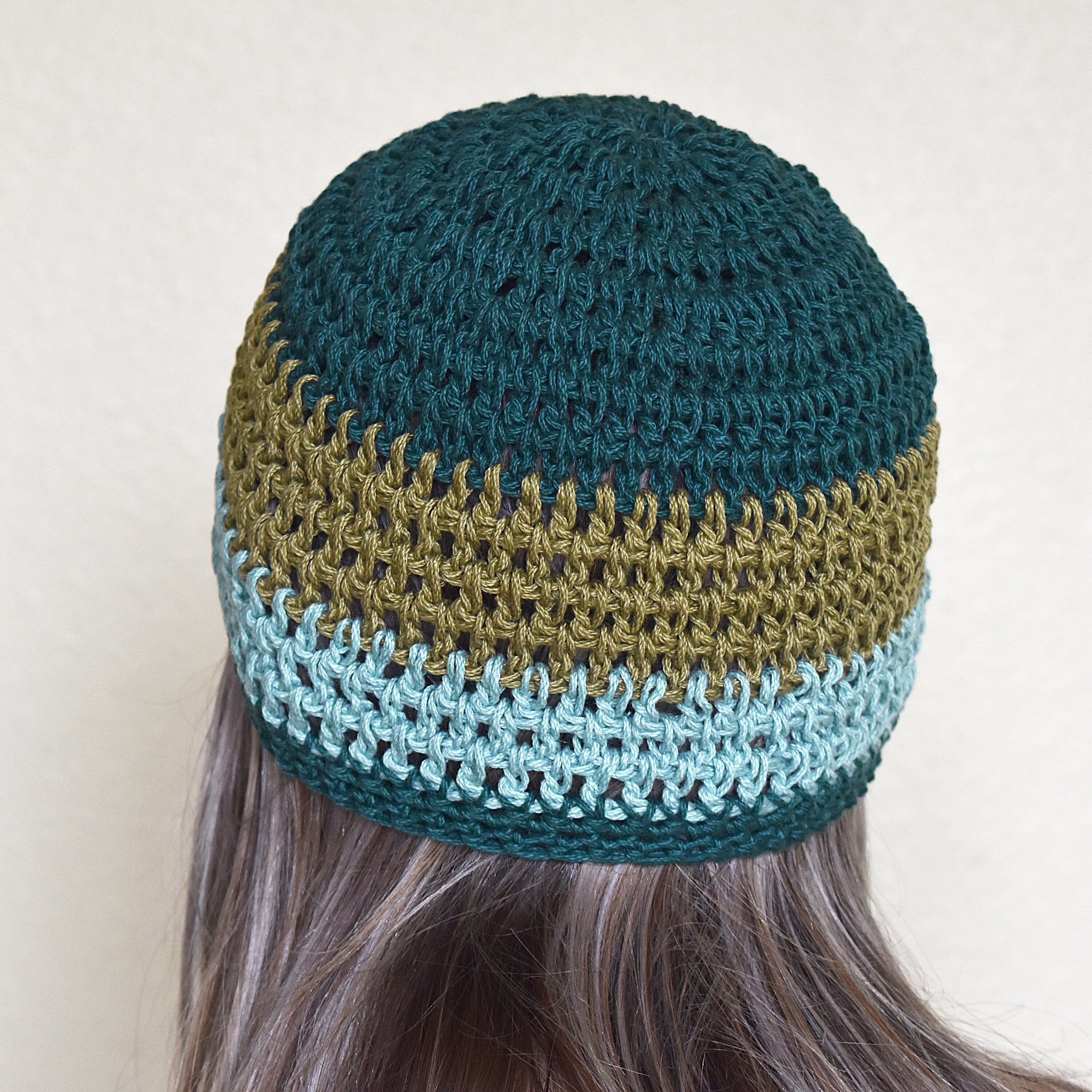 Woman with long hair wearing a crocheted beanie with a teal and green striped pattern against a neutral background.