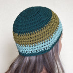 A person wearing a crocheted beanie with a green and teal striped pattern against a neutral background.