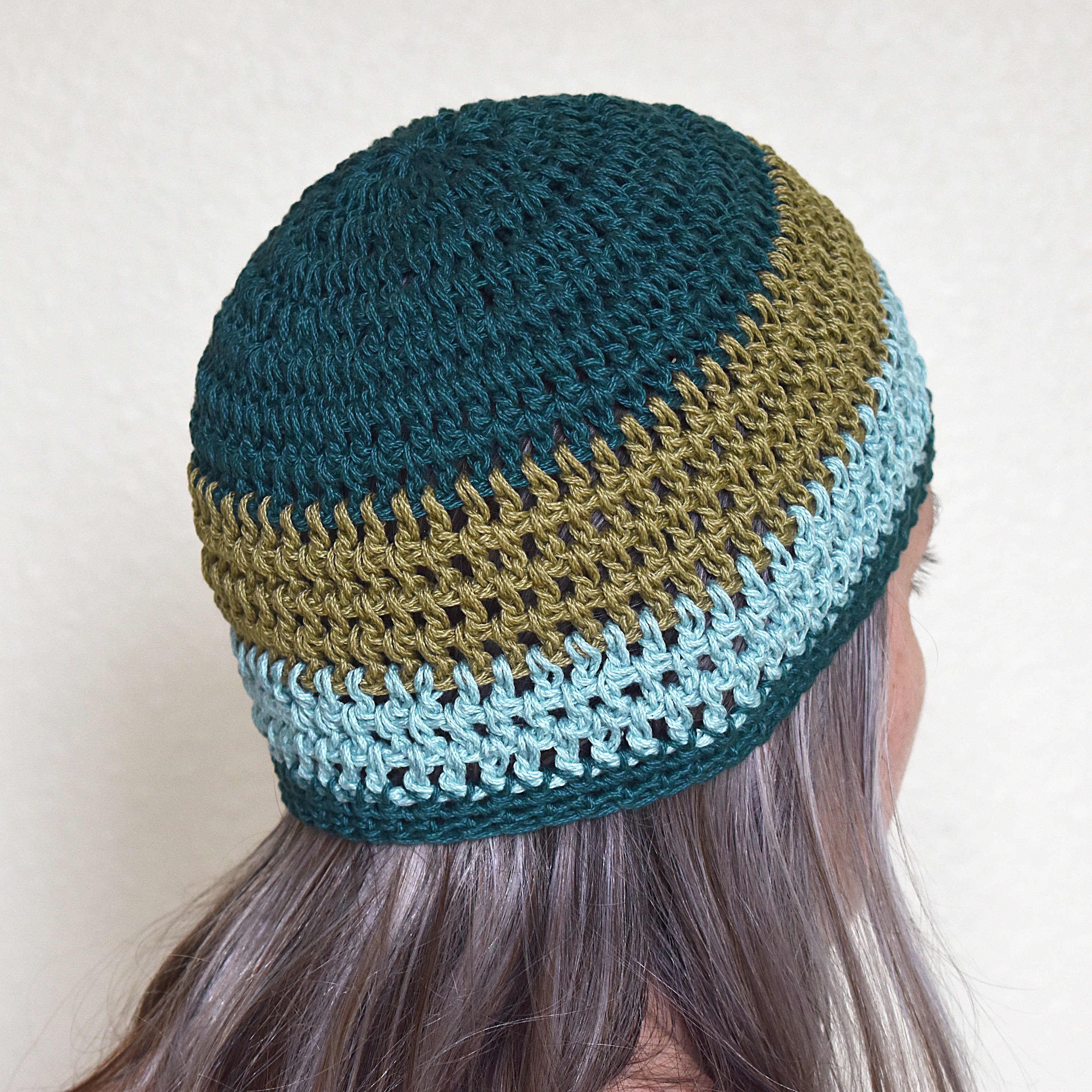 A person wearing a crocheted beanie with a green and teal striped pattern against a neutral background.