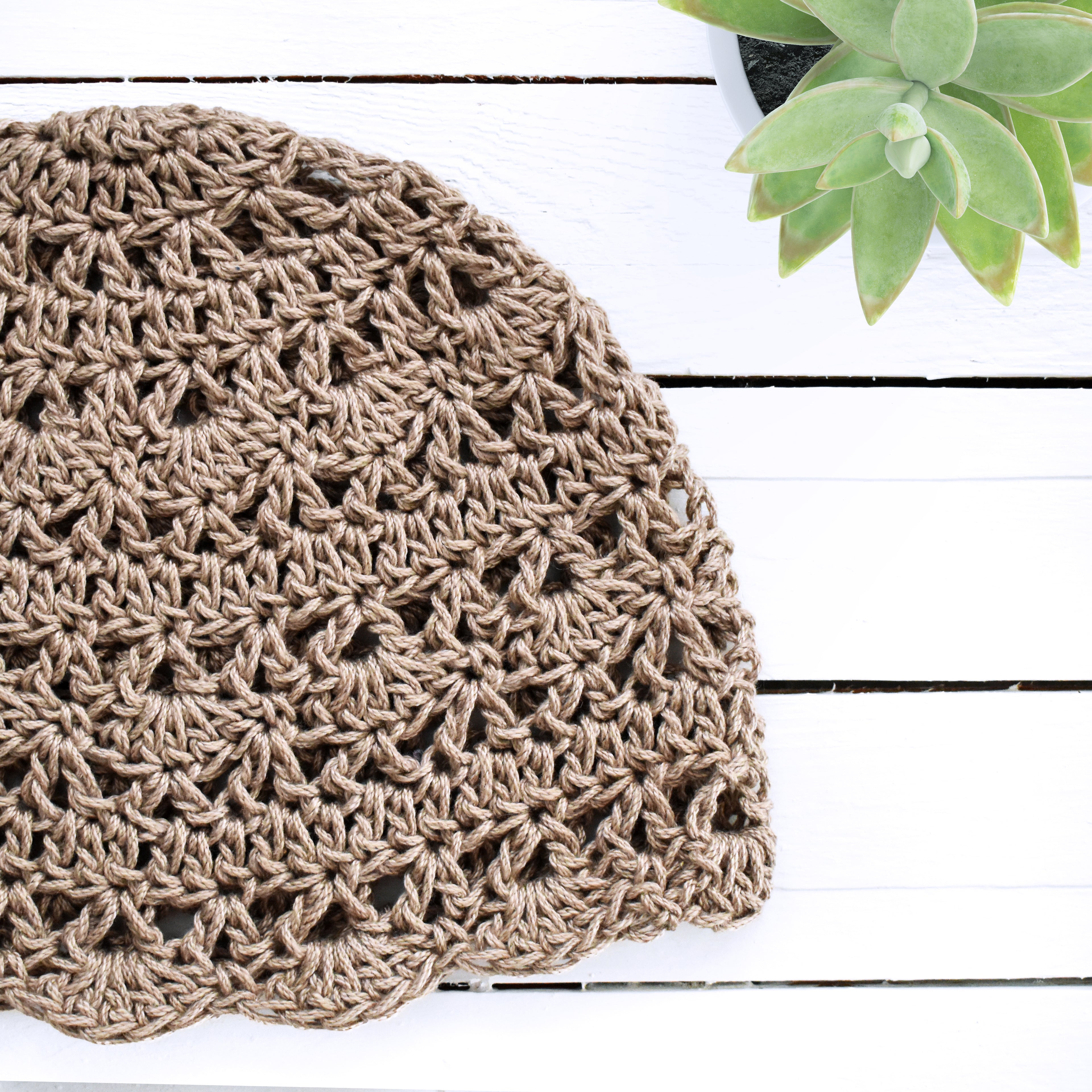 Taupe crochet hat on a white wooden surface with a green plant in the corner.