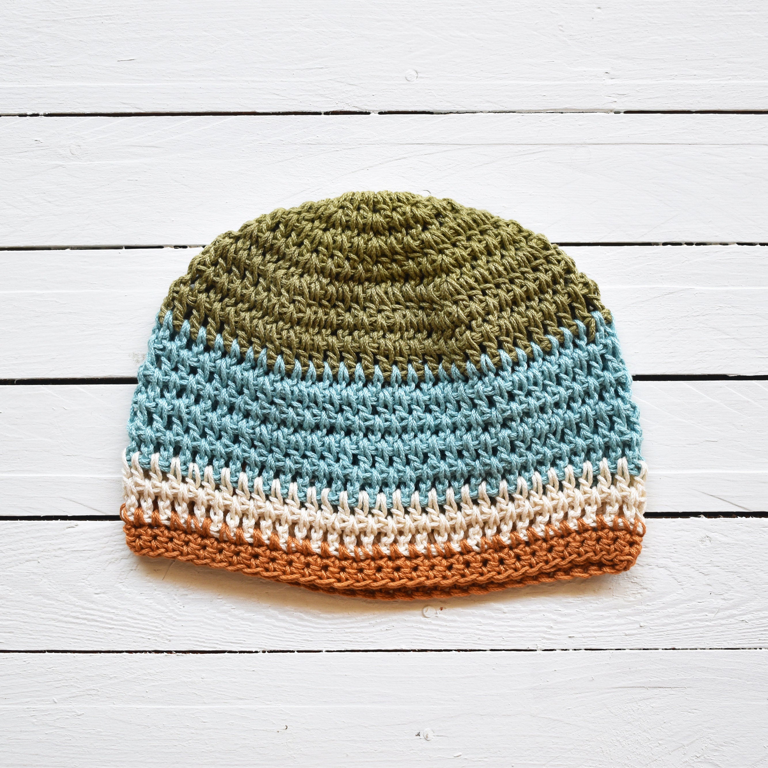 Crocheted beanie with green, light blue, beige, and orange striped colors on a white wooden surface.