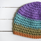 Crocheted beanie with purple, teal, green, and orange striped colors on a white wooden surface.