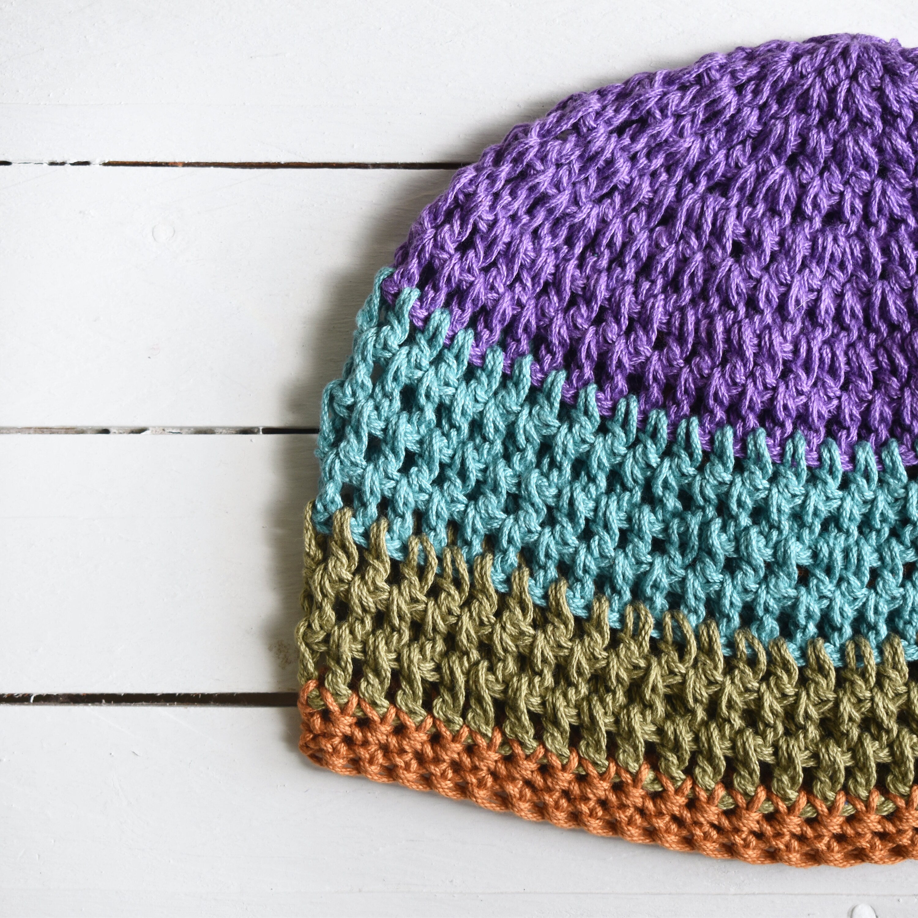 Crocheted beanie with purple, teal, green, and orange striped colors on a white wooden surface.