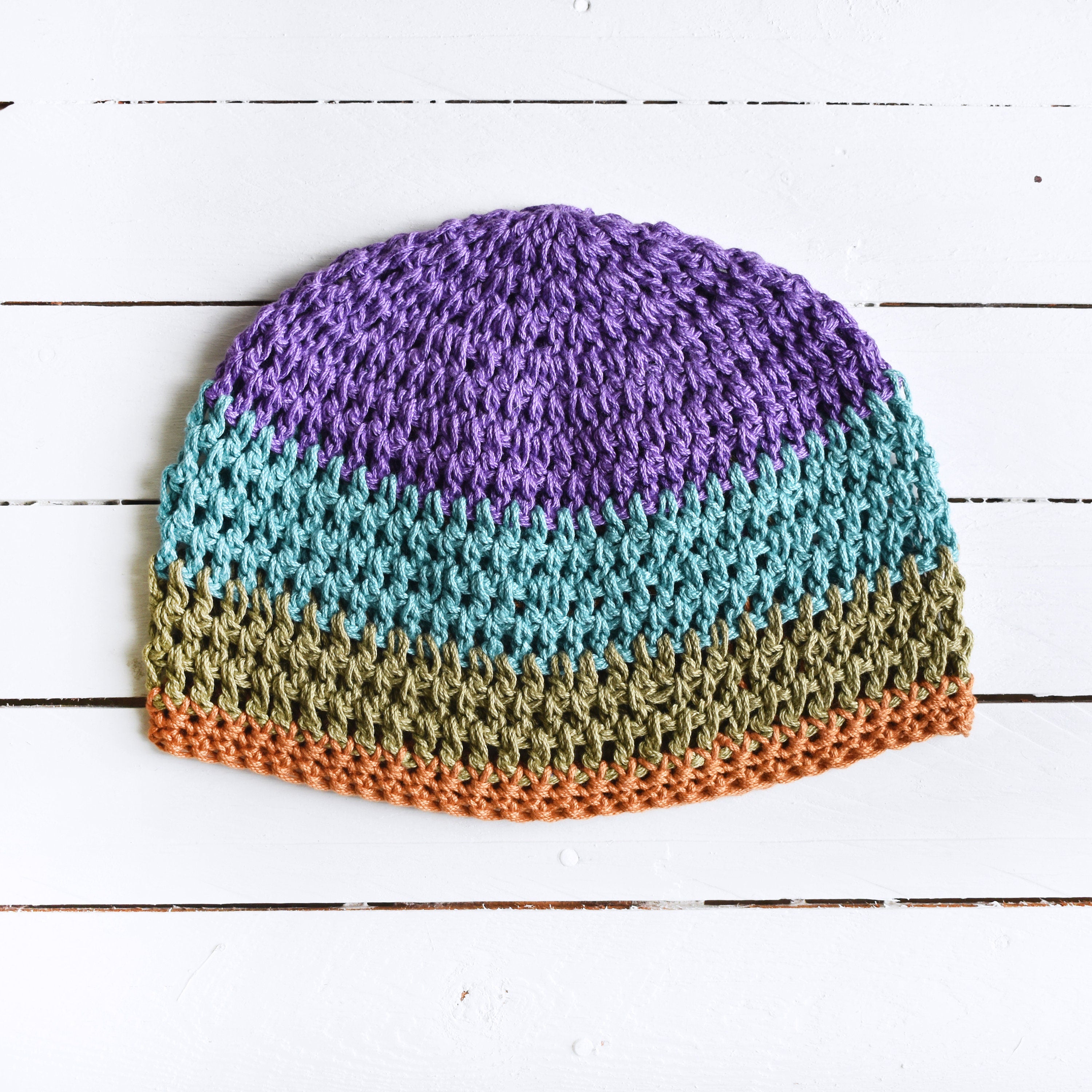 Colorful crocheted beanie with purple, blue, green, and orange stripes on a white wooden surface.