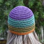 A person wearing a colorful knitted beanie with purple, blue, green, and orange stripes against a blurred natural outdoor background.