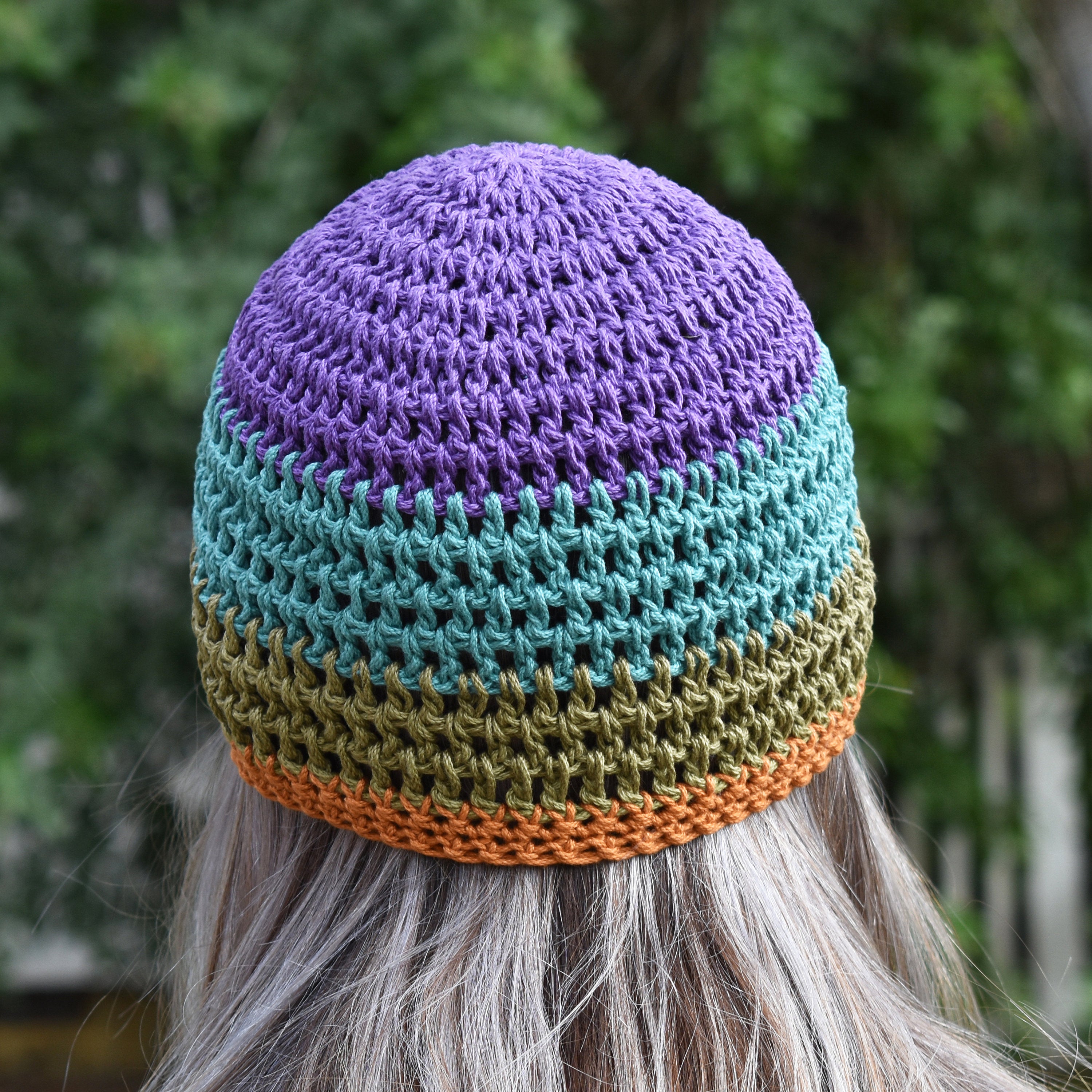 A person wearing a colorful knitted beanie with purple, blue, green, and orange stripes against a blurred natural outdoor background.