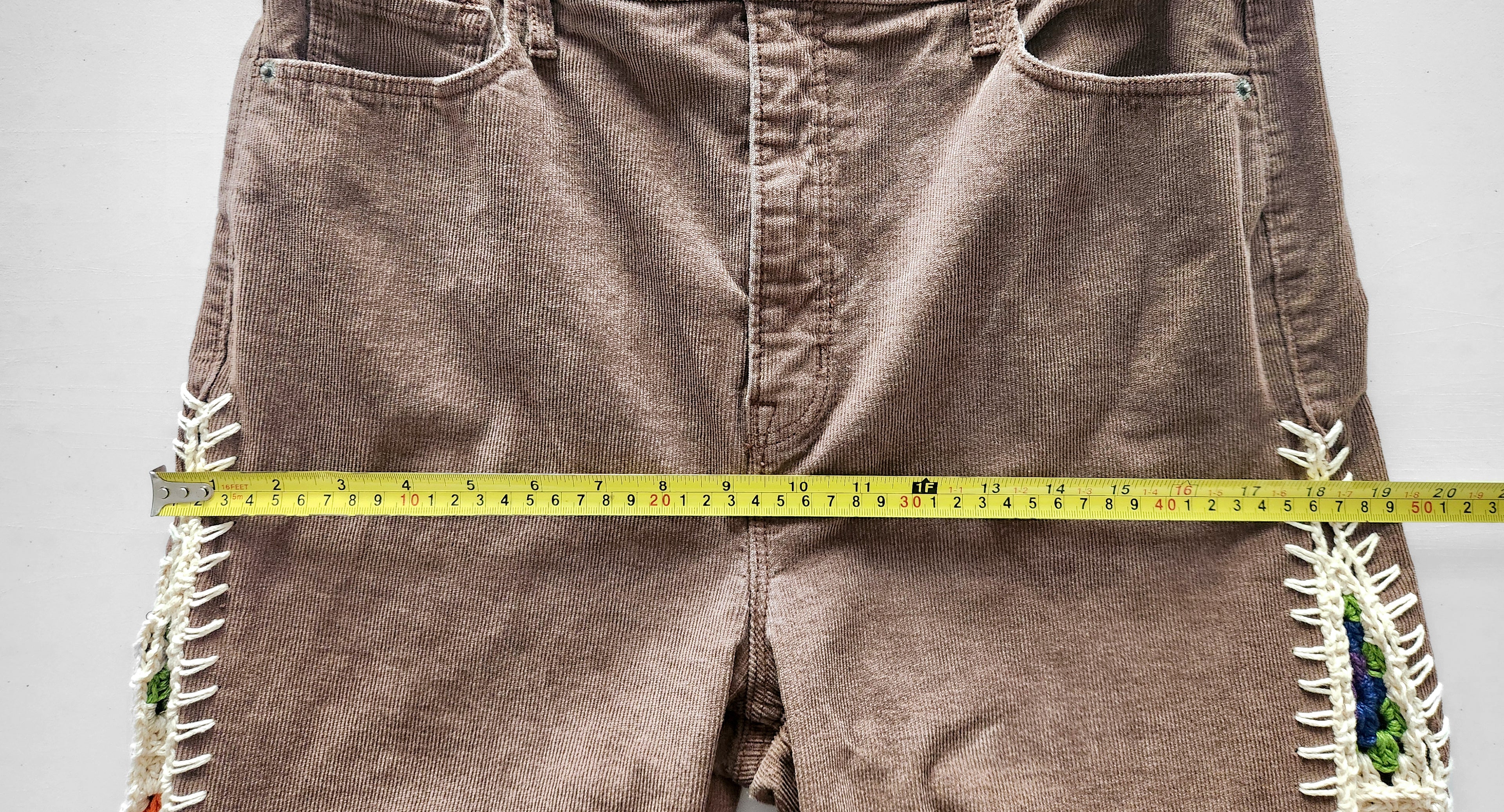 Brown corduroy pants with decorative crochet inserts and a measuring tape across the hips.
