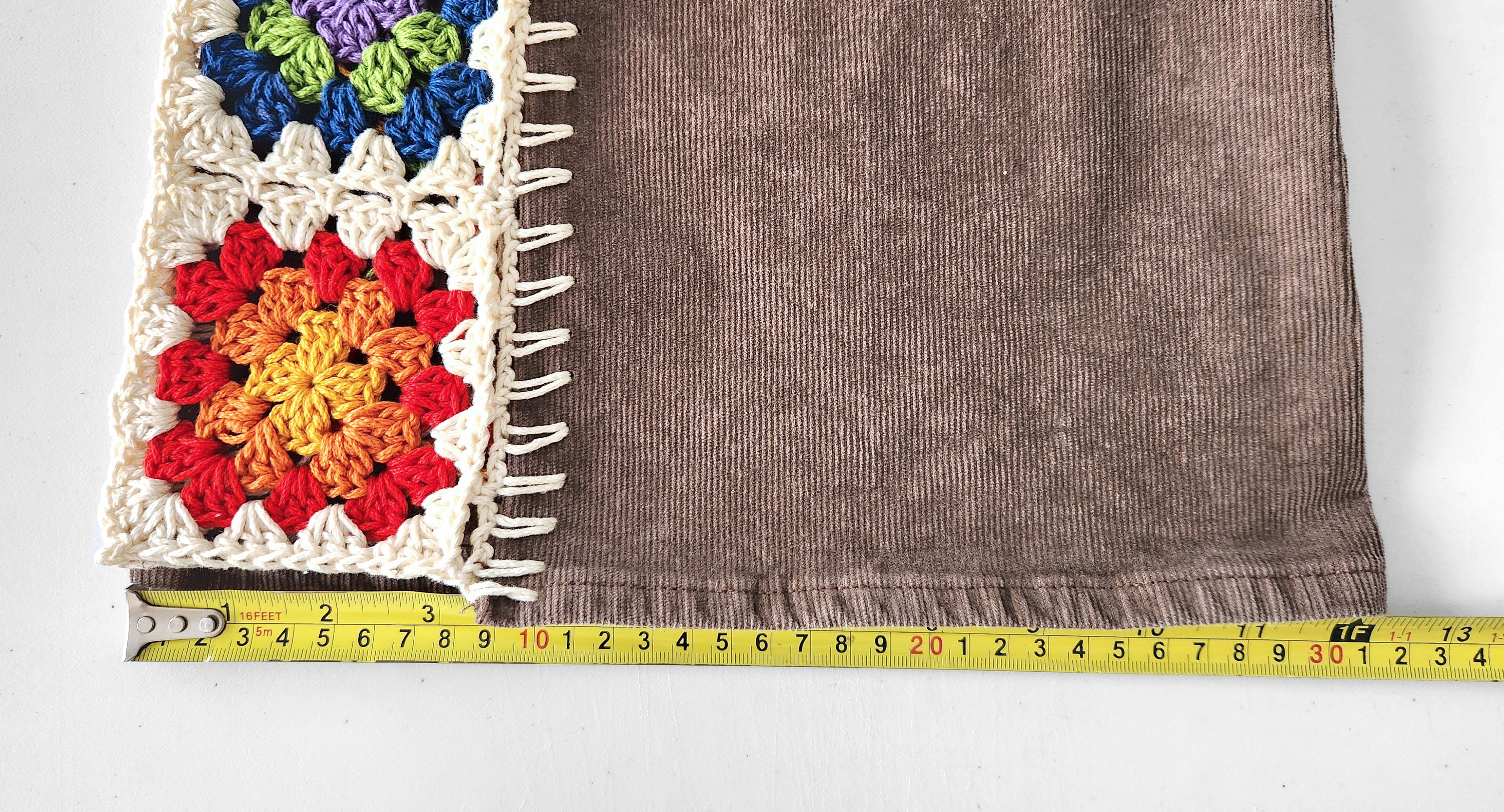 Measuring the leg opening of brown corduroy pants with colorful crochet square inserts.