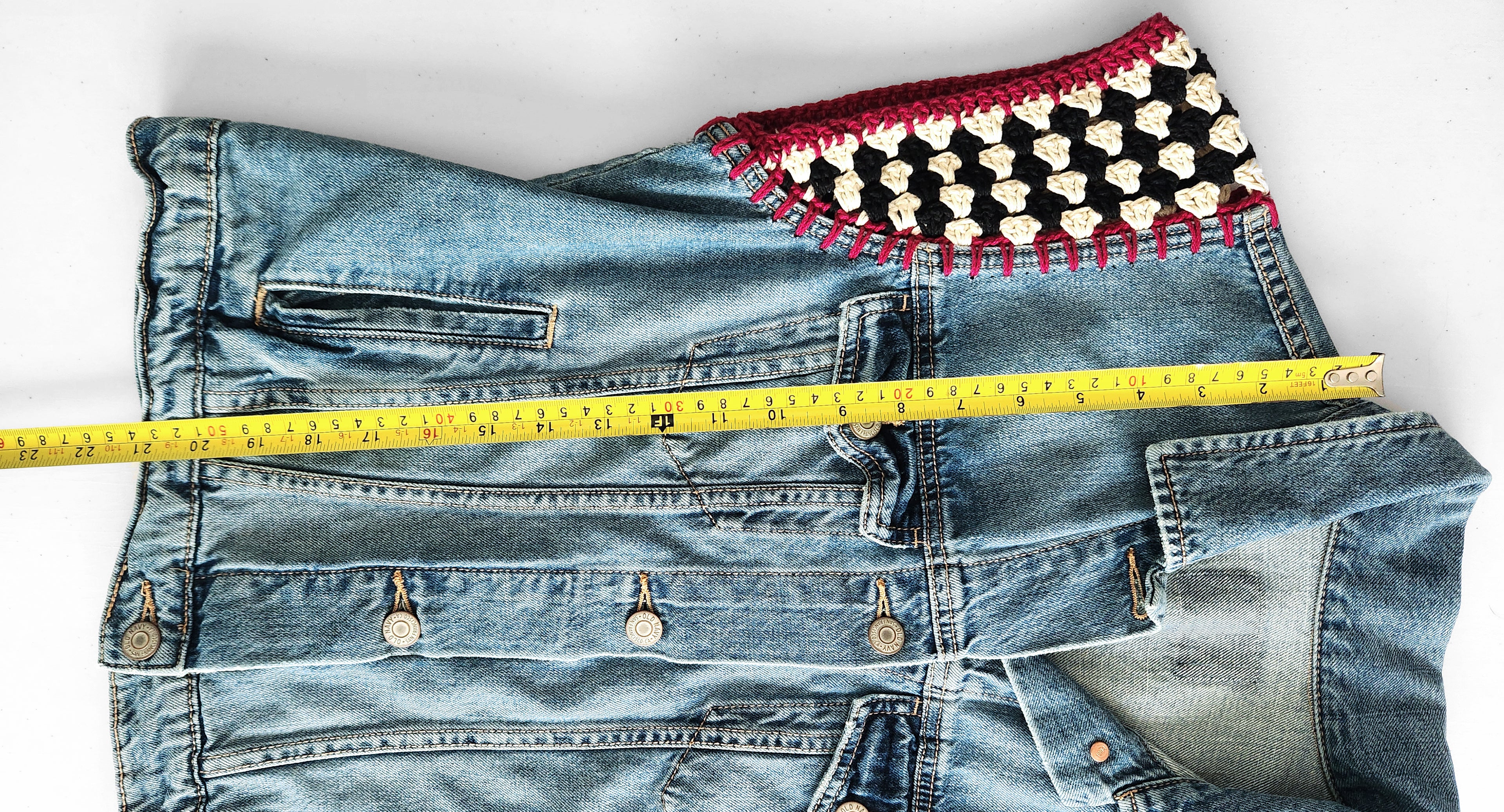 Denim jacket with a colorful crochet cap sleeve and a measuring tape on a white background.