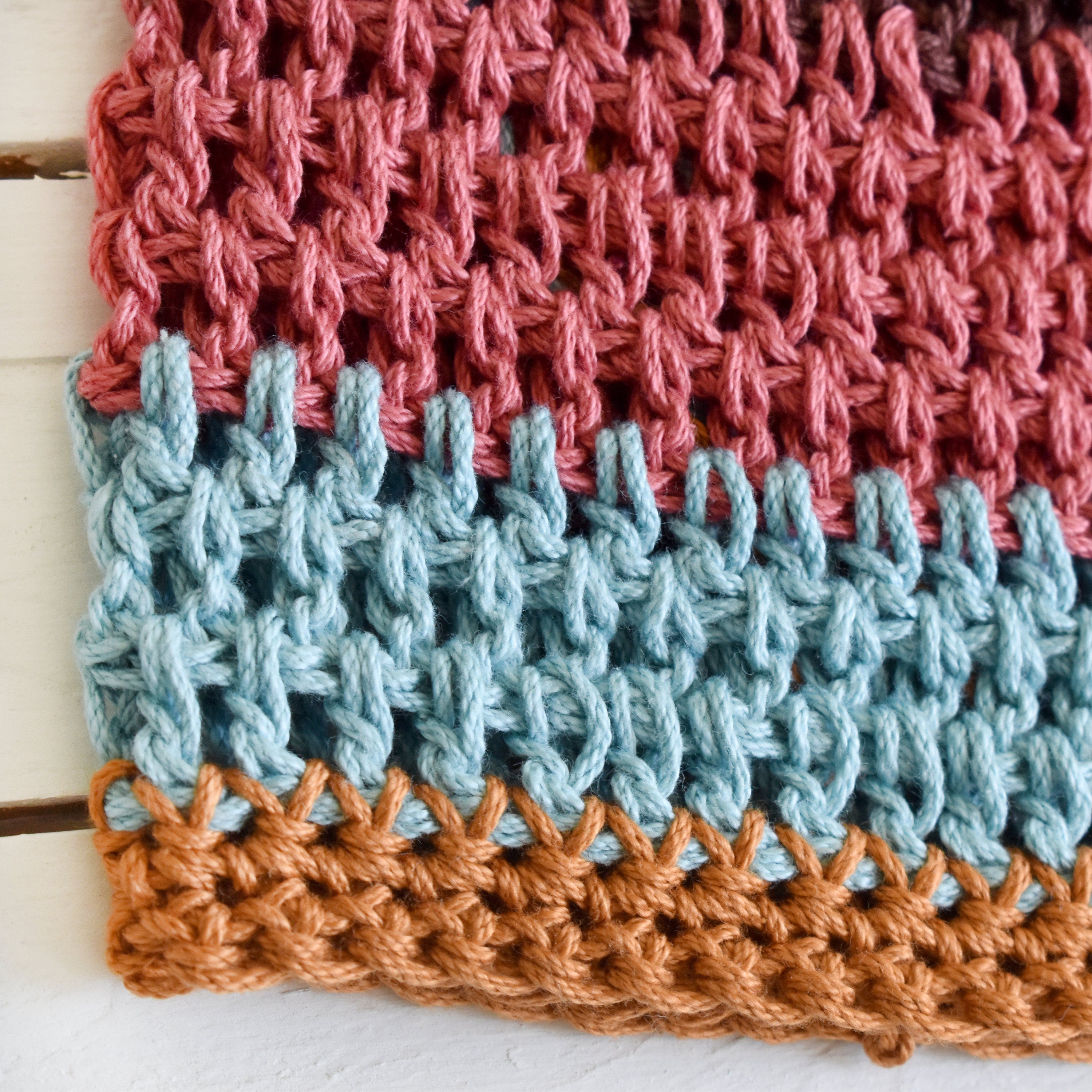 Close-up of a crocheted hat with a striped pattern in terracotta pink, blue, and orange on a wooden surface.