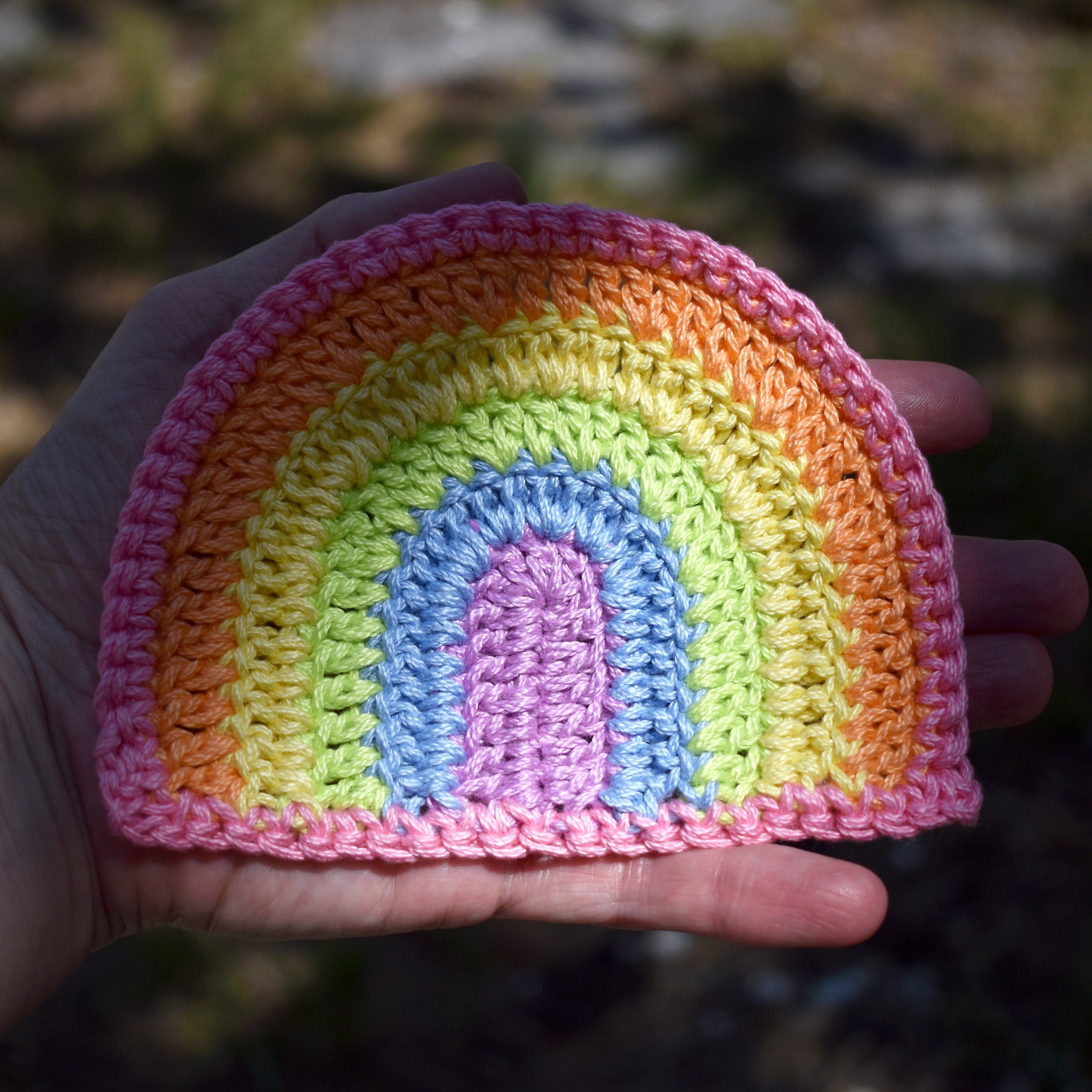 Hand holding a crocheted rainbow patch with pastel colors against a blurred natural background.