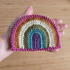 Hand holding a crocheted rainbow patch on a wooden surface.