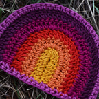 Crocheted rainbow patch with a gradient of purple, pink, orange, and yellow on grass.