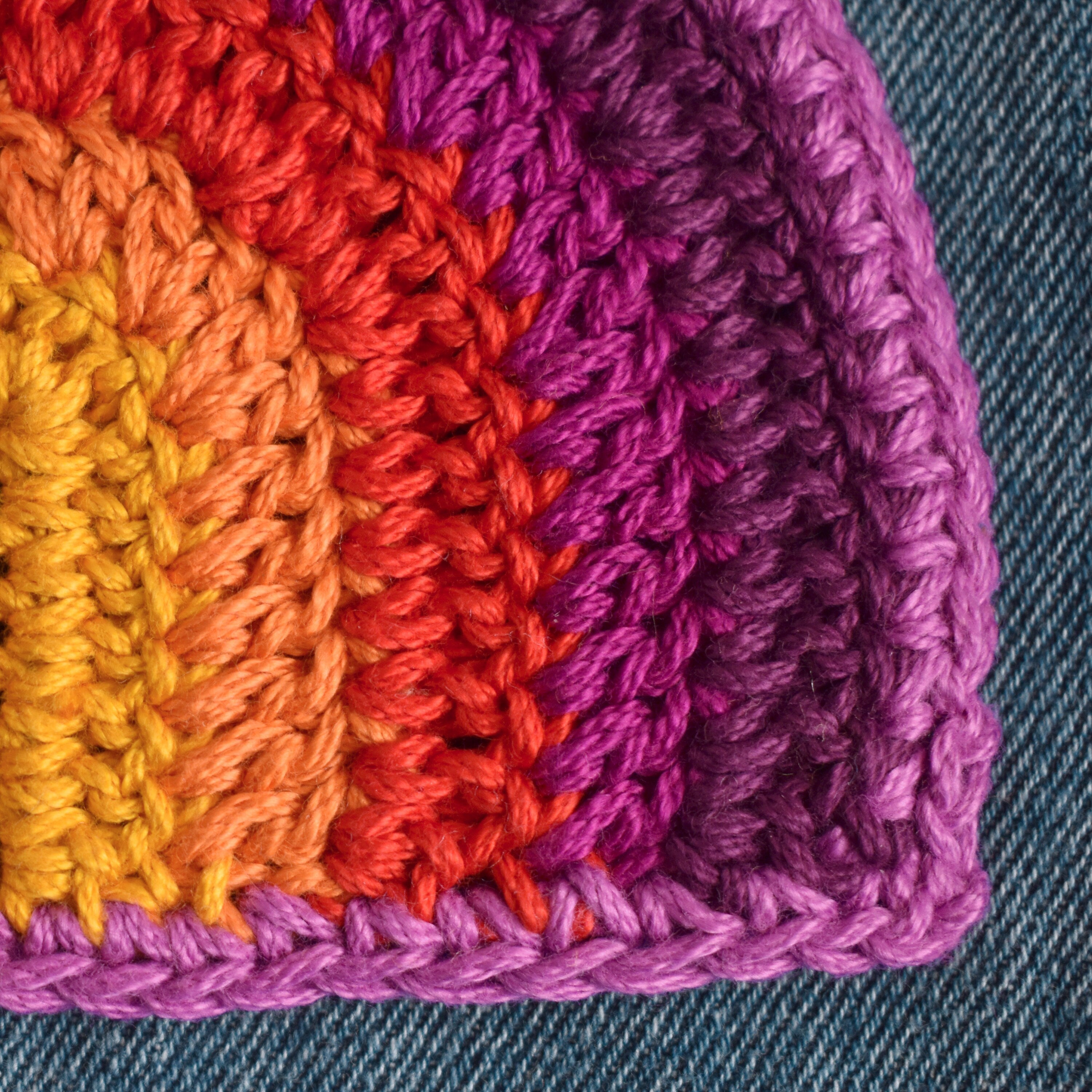 Close-up of a colorful crocheted corner with orange, red, and purple yarn on a denim background.