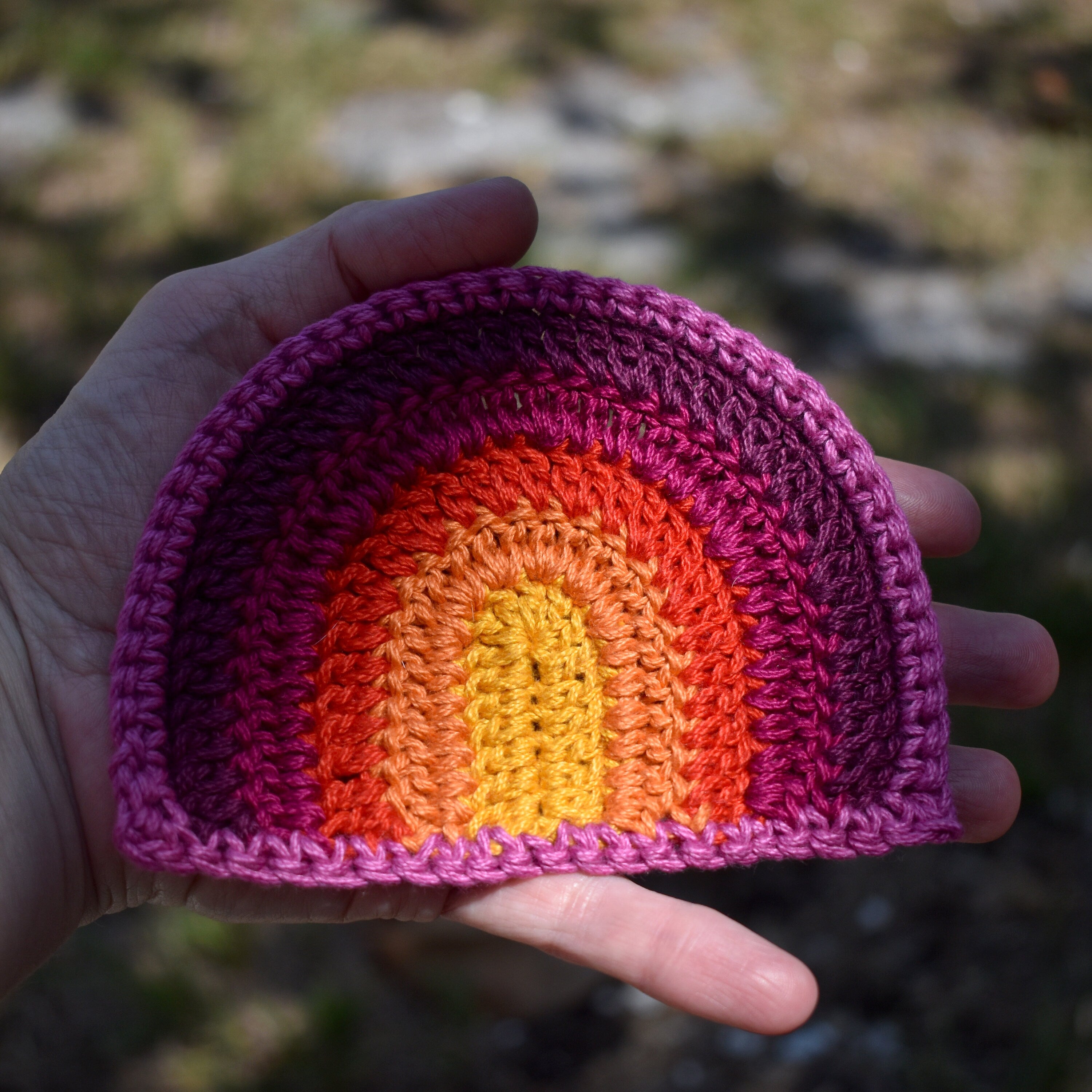 Hand holding a crocheted rainbow-patterned item with a blurred natural background.
