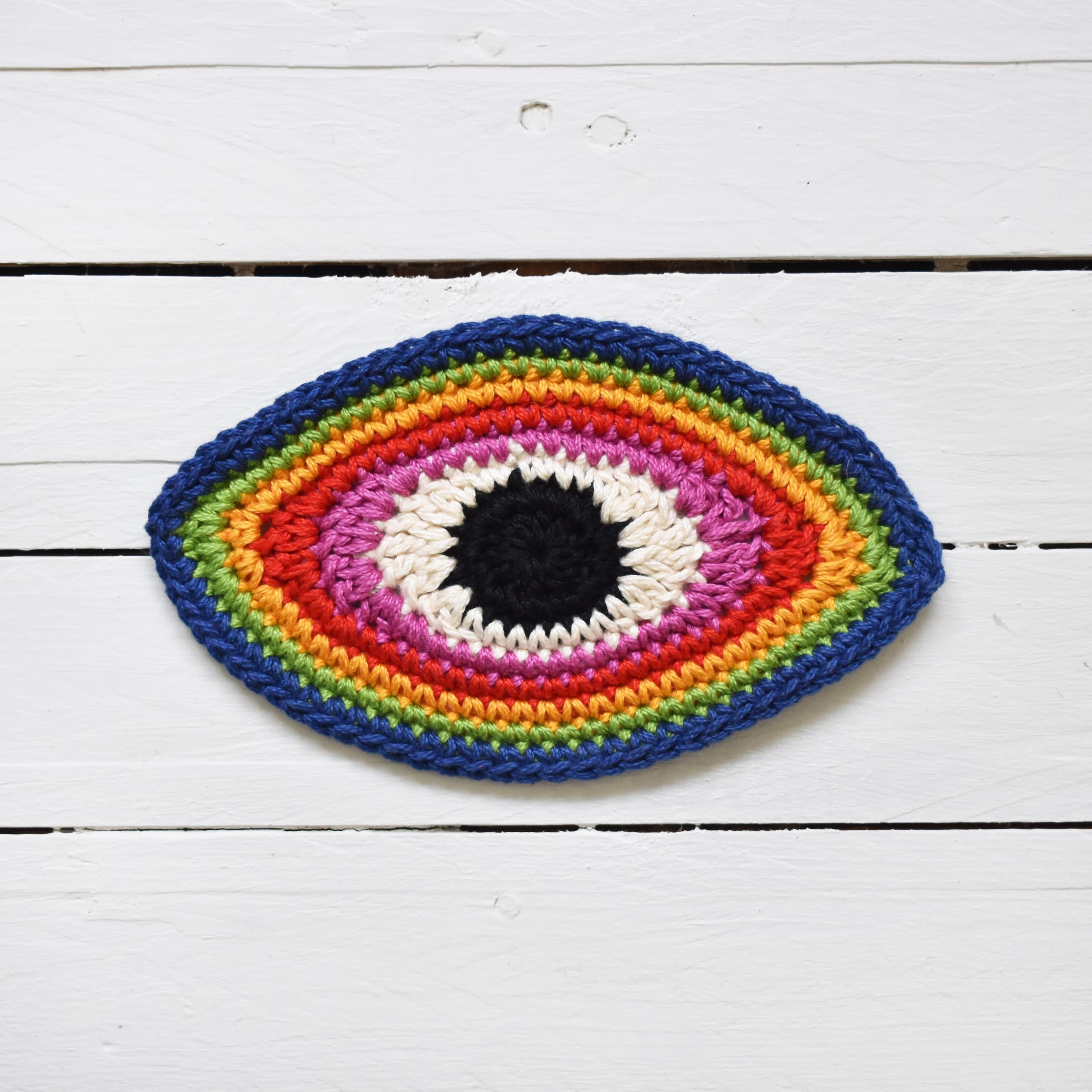 Crocheted multicolored eye-shaped patch on a white wooden surface.