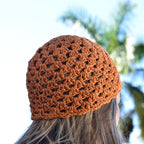An orange crochet hat is worn by a woman looking into a blurred natural background.