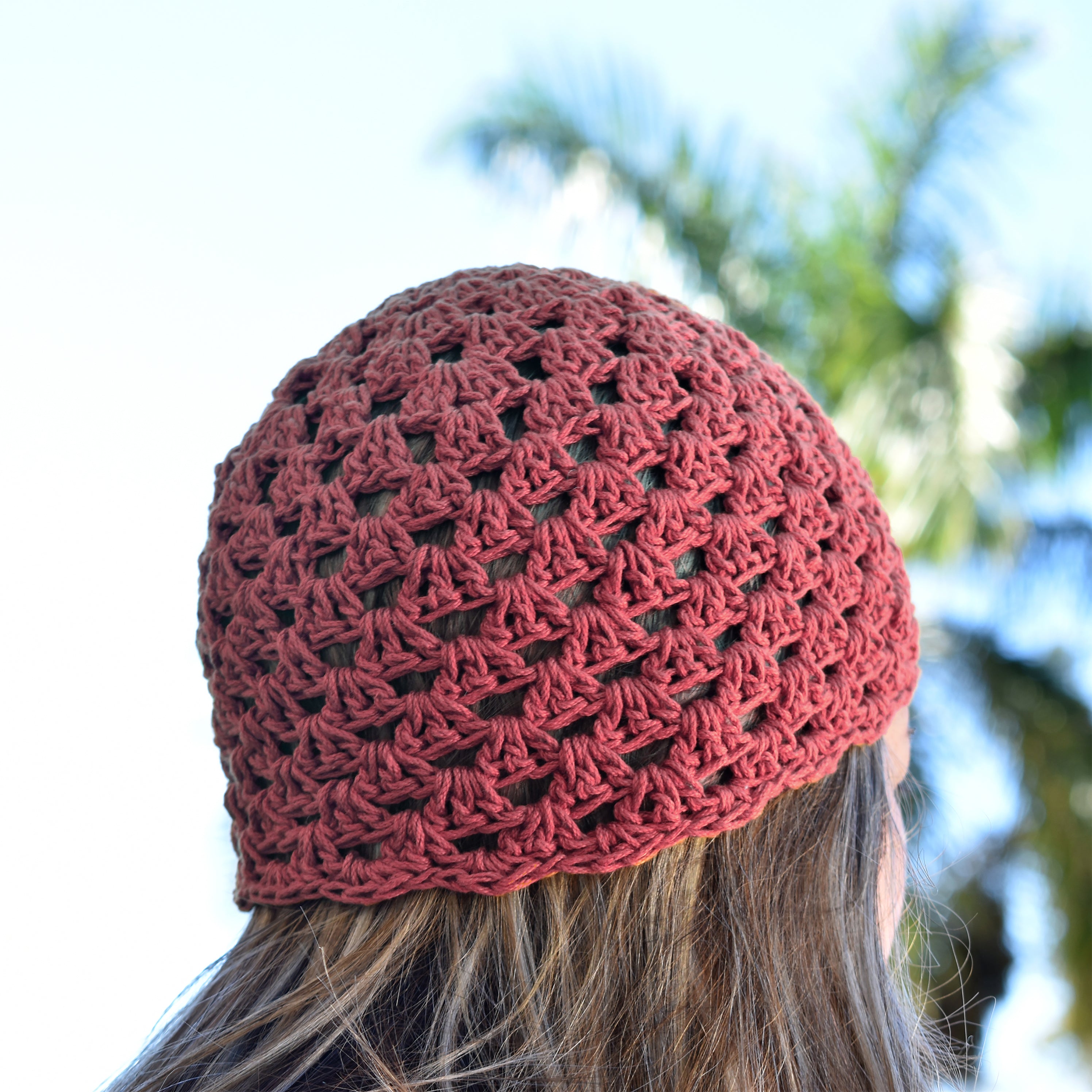 Terracotta pink crochet cap worn by a person with blurred greenery in the background.