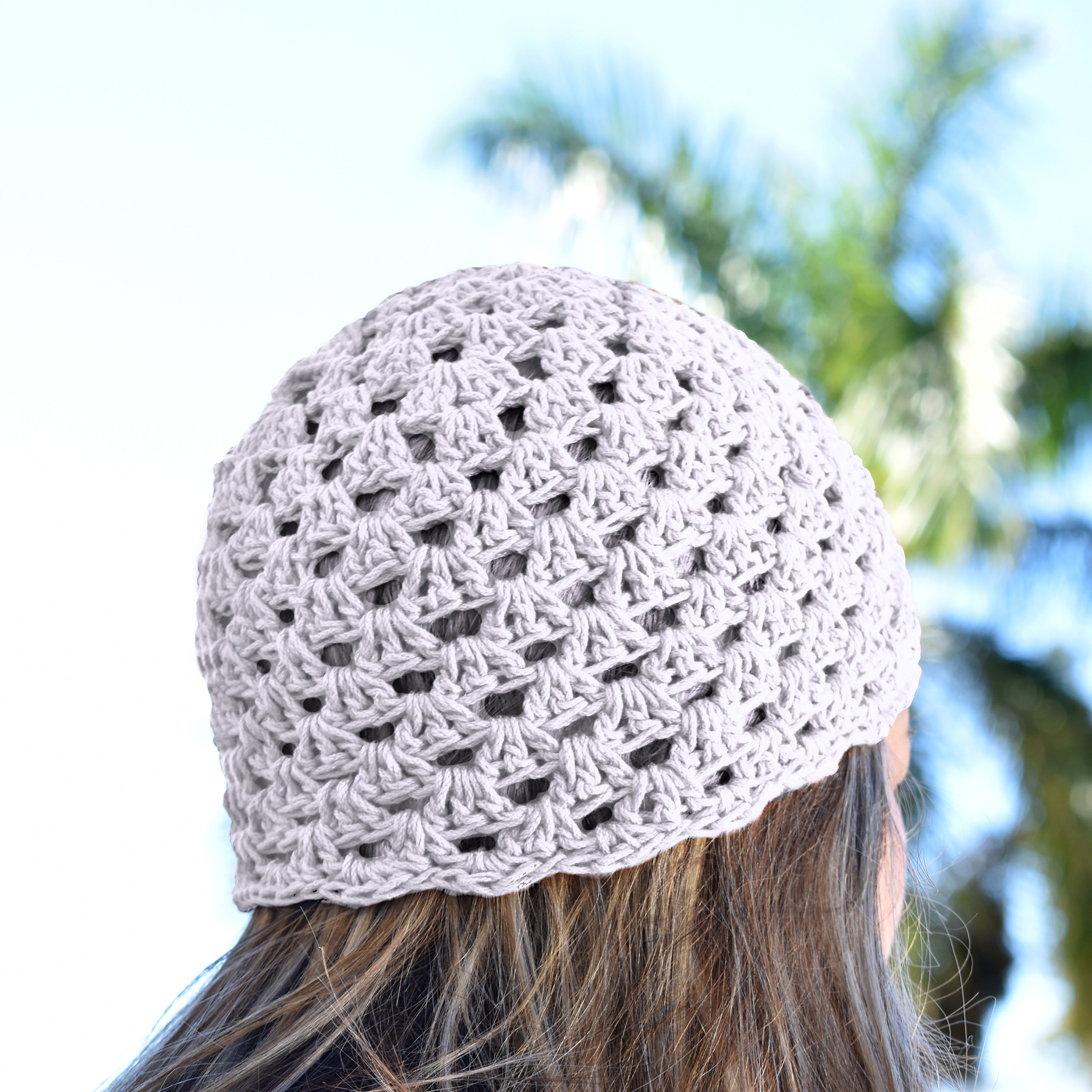 White crocheted beanie worn by a person with blurred greenery in the background.