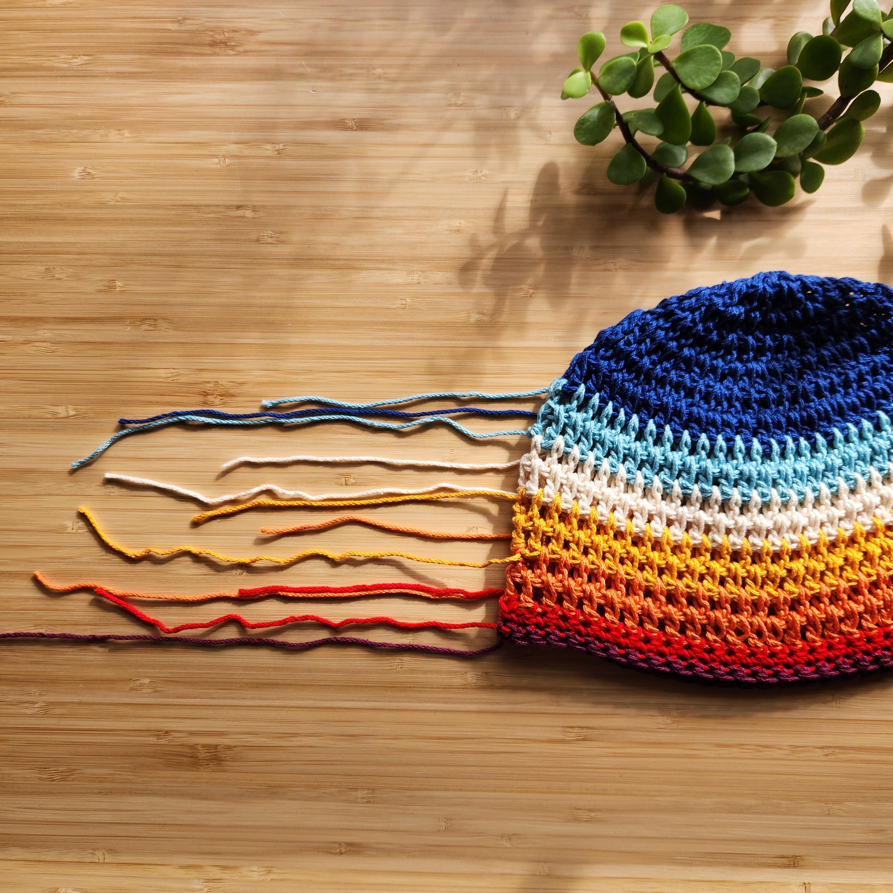 A colorful striped cotton crochet hat on a bamboo table with unfinished, loose strands of yarn stretched out to the side.