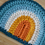 Close-up of a crocheted rainbow with blue, white, orange, and yellow on a wooden surface.