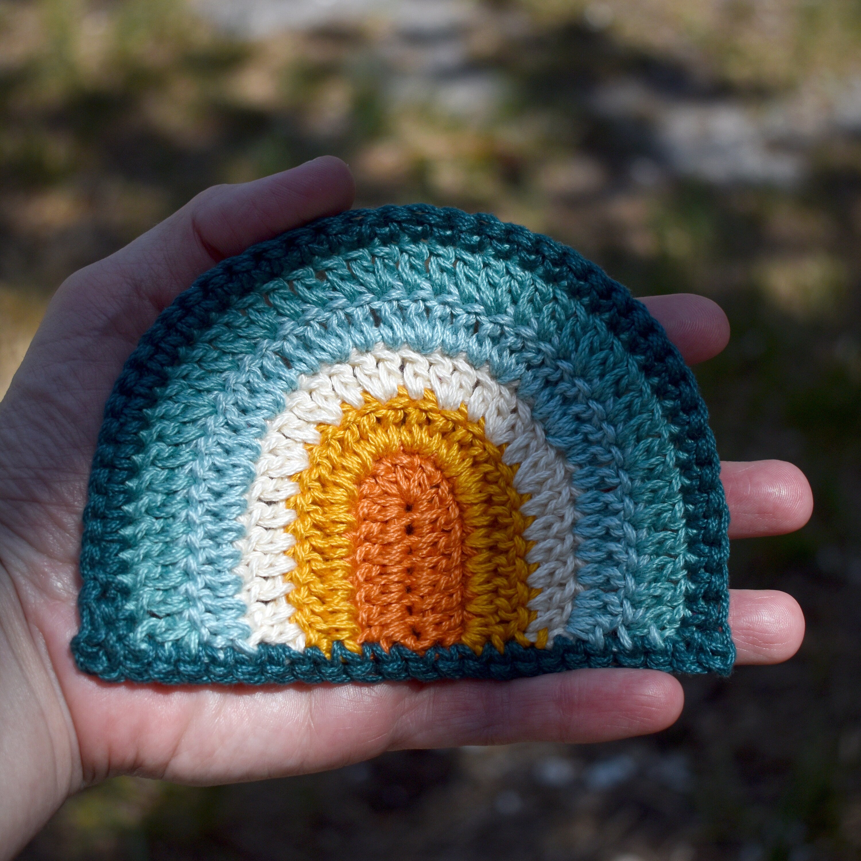 Hand holding a crocheted rainbow patch with a colorful design against a natural background.