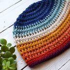 Colorful crocheted hat on a white wooden surface with a small plant.