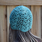 Teal crocheted beanie worn by a person with long hair, against a wooden fence background.